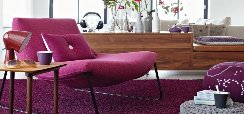 Ein stilvolles Wohnzimmer mit einem leuchtend rosa Sessel, einem hölzernen Sideboard und dekorativen Akzenten auf einem violetten Teppichboden.