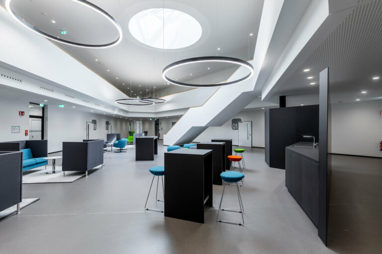 Moderne Bürolounge mit runden Leuchten, geometrischen Möbeln in verschiedenen Farben und einer zentralen Treppe mit Linoleumboden.