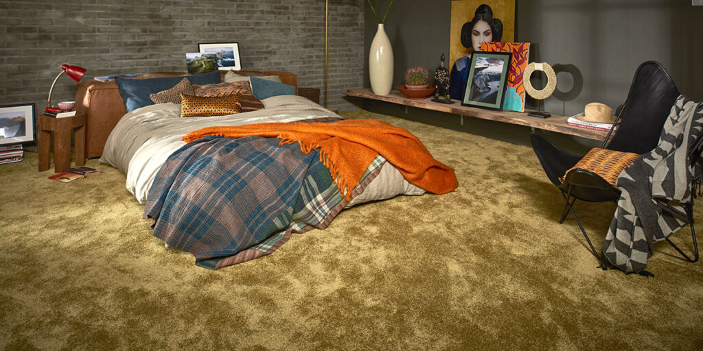 Modernes Schlafzimmer mit einem Bett mit karierter Bettwäsche, einer orangefarbenen Überwurfdecke, schwebenden Regalen mit Kunstwerken und Fotos sowie einem schwarzen Stuhl in einem Raum mit Backsteinwänden und Teppichboden.
