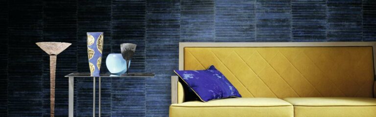 Bild Vliestapete - Ein modernes Wohnzimmer mit einer gelben Couch mit blauen Kissen, einem Metall-Beistelltisch mit dekorativen Vasen und einer Lampe vor einer Wand tapeziert mit blauer Vliestapete.
