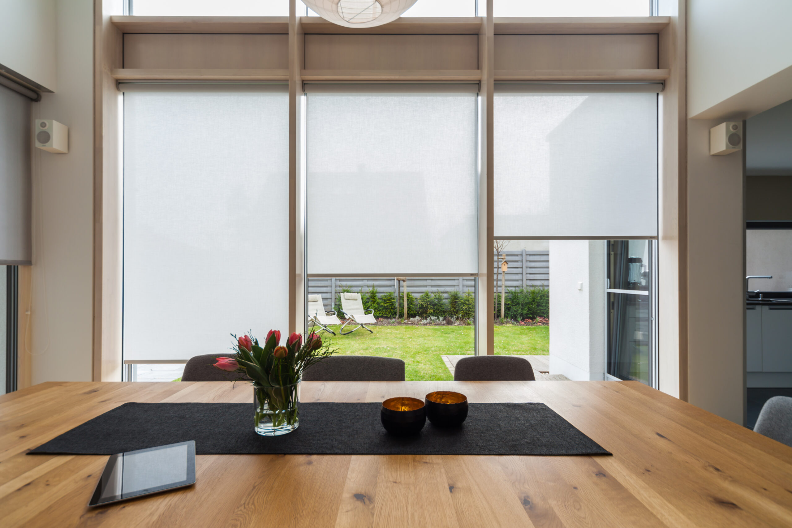 Modernes Esszimmer mit einem großen Tisch, zwei Schüsseln und einem Blumenstrauß und Blick auf einen Hinterhof durch raumhohe Fenster.