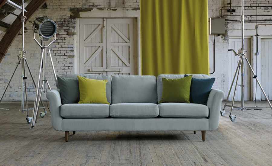Modernes Wohnzimmer mit einem Sofa mit grauem Polsterstoff, geschmückt mit grünen und gelben Kissen, Strahlern und einem passenden Vorhang, vor einer rustikalen weißen Ziegelwand mit Vinylboden.