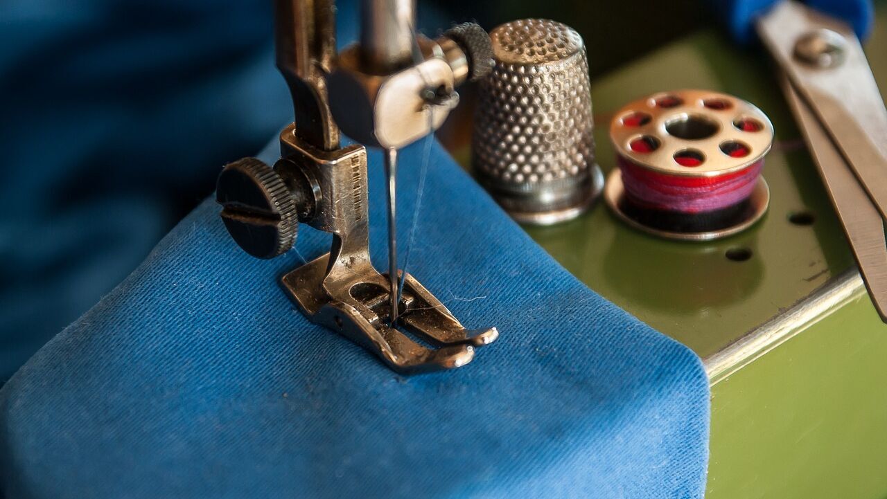 Gardinennäherei - Nähservice -Nahaufnahme der Nadel und des Nähfußes einer Nähmaschine auf blauem Stoff, mit Schere, Fingerhut und Garnrollen in der Nähe. T&K WOHNSTDIO - Raumausstattung, Bodenbeläge, Vinylboden, Parkett, Gardinen - Saarburger Straße 25, 67071 Ludwigshafen