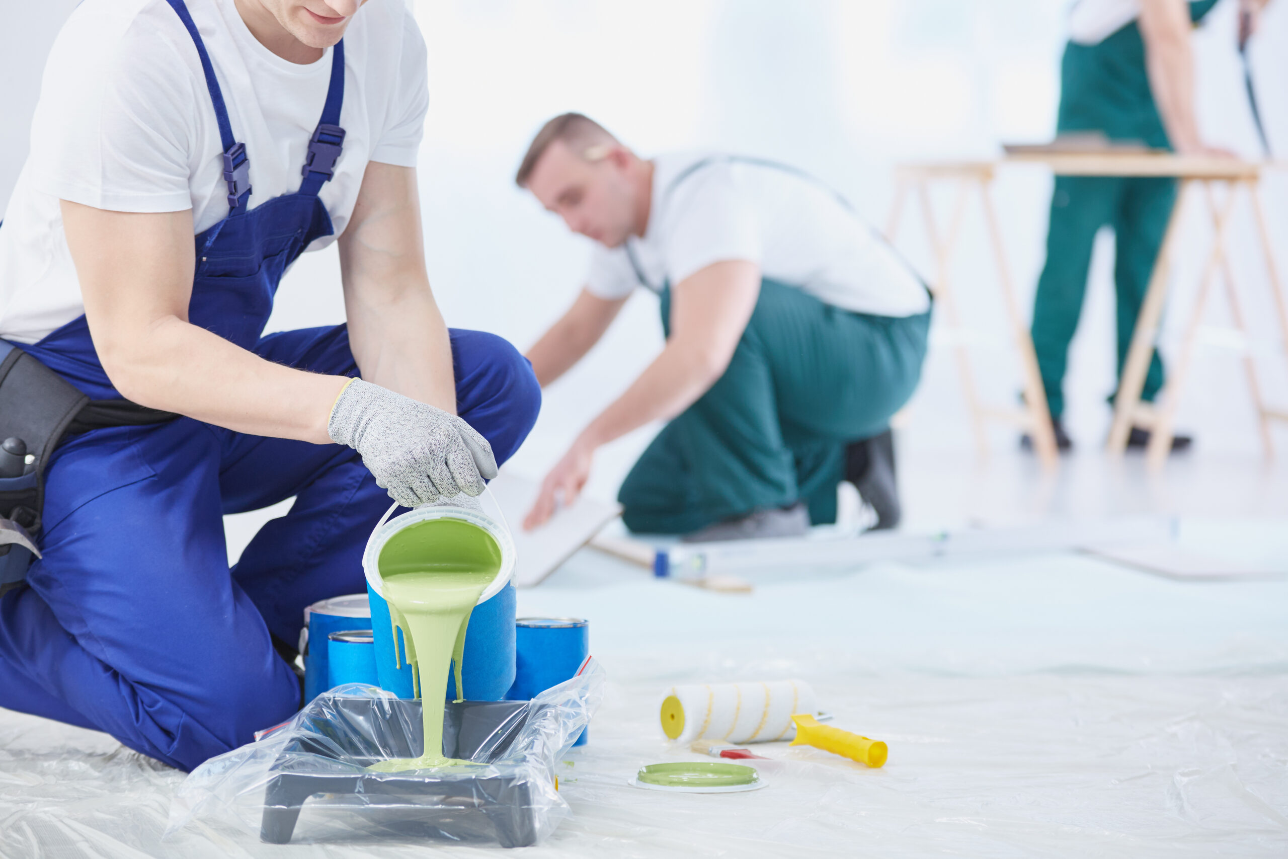 Ein Arbeiter im blauen Overall gießt grüne Farbe auf eine Tablette, während im Hintergrund zwei andere Arbeiter einen Holzfußboden verlegen und schneiden.