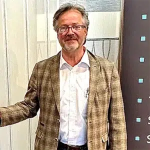 Ein bärtiger Mann mit Brille, bekleidet mit einer karierten Jacke und einem weißen Hemd, steht selbstbewusst vor einem braunen Hintergrund mit blauen Quadraten und verkörpert die Essenz von „Über Uns“. - T&K WOHNSTDIO - Raumausstattung, Bodenbeläge, Vinylboden, Parkett, Gardinen - Saarburger Straße 25, 67071 Ludwigshafen